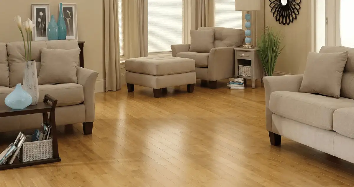 A living room with professionally installed bamboo flooring, beige sofas, a matching ottoman, a side table with a blue lamp, vases, framed pictures, and large windows with blinds and curtains in Oklahoma City, OK
